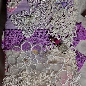 Può includere: Una copertina per diario in tessuto rettangolare con base color lavanda, decorata con applicazioni in pizzo color crema che includono motivi a farfalla e floreali. Il design presenta un bordo all'uncinetto viola, piccole perle e una spilla decorativa.
