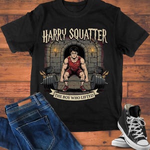 Op de afbeelding: Zwart T-shirt met een afbeelding van een gewichtheffer in rood die een halter optilt. De tekst "HARRY SQUATTER" staat erboven en "THE BOY WHO LIFTED" eronder. Gecombineerd met een blauwe jeans en zwarte hoge sneakers.