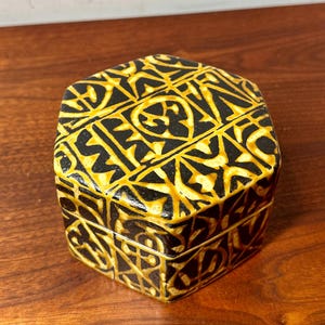 Puede incluir: Una caja decorativa hexagonal negra y dorada con tapa. La caja presenta un patrón geométrico repetitivo en dorado sobre un fondo negro. La caja está sobre una superficie de madera.