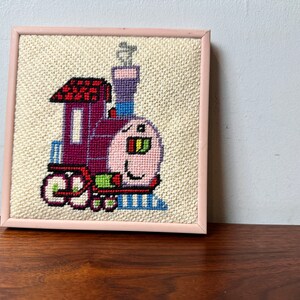 May include: A framed cross-stitch of a cartoon train with a pink face, red roof, and blue and green details. The train is on a white background.