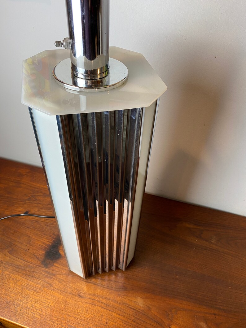 Op de afbeelding: Een chromen en witte tafellamp met een geometrische basis en een geribbeld ontwerp. De lamp heeft een witte kap en een chromen steel.