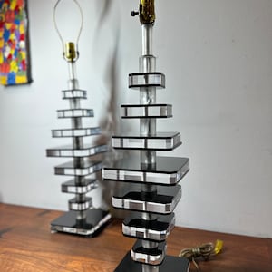 May include: Two modern table lamps with a stacked, geometric design. Each lamp features a black square base, a silver metal central column, and multiple clear, rectangular tiers. The lamps are on a wooden surface.