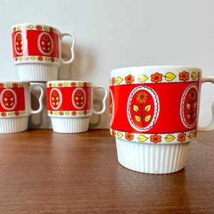 May include: Set of four vintage stacking mugs. Each mug features a white base and handle, with a vibrant red band adorned with floral patterns. The design includes a central oval with a flower and leaf motif.