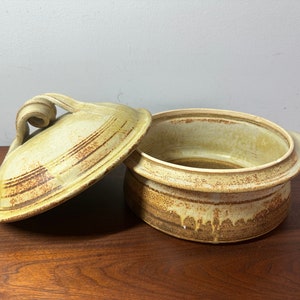 May include: A ceramic casserole dish with a lid. The dish is a light brown colour with a glaze that has a crackled effect. The lid has a unique handle that is shaped like a swirl.