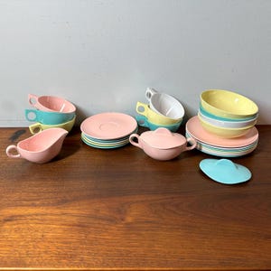 Pode incluir: Uma coleção de louças vintage em cores pastel, incluindo xícaras, pires, tigelas, um molheiro e uma açucareira com tampa. O conjunto apresenta peças rosa, amarelas, azuis e brancas, empilhadas e dispostas sobre uma superfície de madeira. Uma tampa azul clara também está presente.