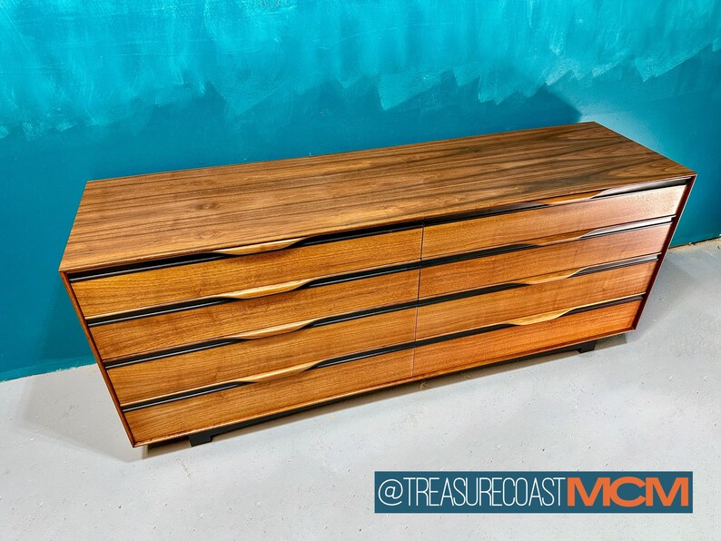 May include: A six-drawer dresser made of wood with a dark brown finish. The drawers have black trim and wooden handles. The dresser is sitting on a light gray floor.