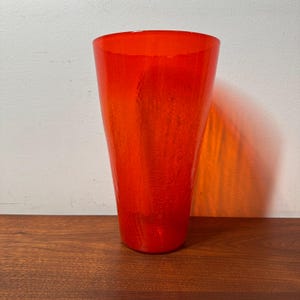 May include: A tall, vibrant orange glass vase with a textured surface. The vase has a conical shape, wider at the top and tapering towards the base. The vase is displayed on a dark wooden surface.