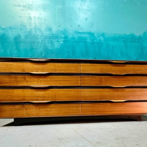 May include: A six-drawer dresser with a mid-century modern design. The dresser is made of wood with a light brown finish and has black accents. The drawers have brass handles.