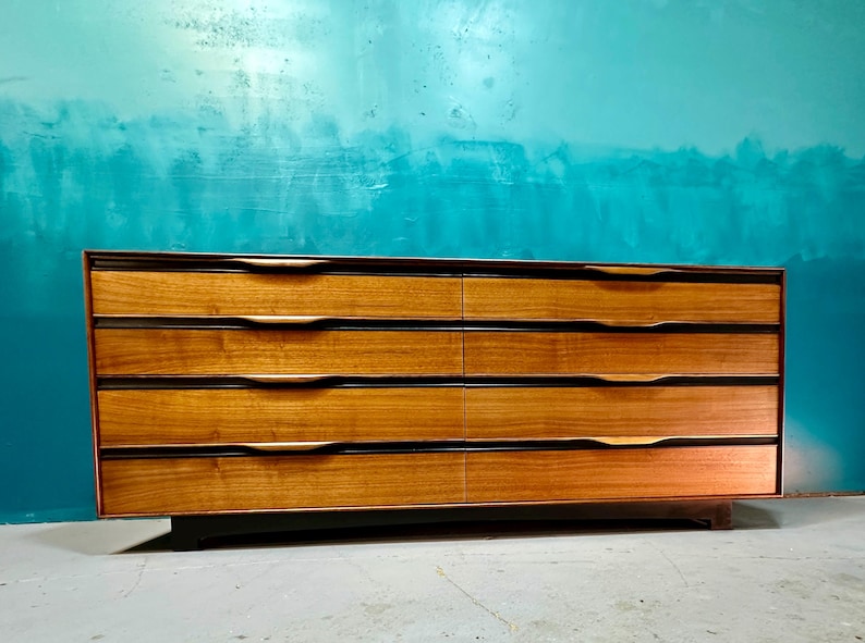 May include: A six-drawer dresser with a mid-century modern design. The dresser is made of wood with a light brown finish and has black accents. The drawers have brass handles.