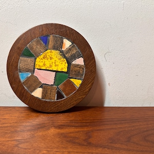Pode incluir: Tábua redonda de madeira com um design de mosaico. O mosaico apresenta um padrão de sol com azulejos amarelos, rosa, verdes e azuis, rodeados por peças de madeira castanha. A tábua está sobre uma superfície de madeira.