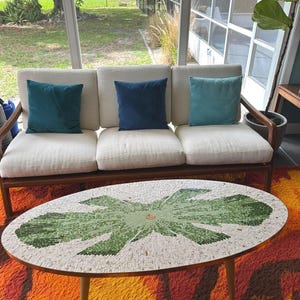 May include: A mid-century modern living room set with a cream-colored sofa, three teal throw pillows, and a mosaic-tiled coffee table. The table has a green and white floral design. The room is set on a colorful orange and brown shag rug.
