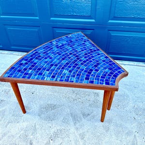 May include: A triangular side table with a blue mosaic tile top and three wooden legs. The table is made of wood and has a natural finish.