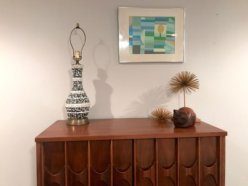 May include: A black and white ceramic lamp with a gold base sits on a dark wood dresser with a geometric abstract painting on the wall behind it.