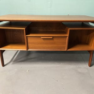 May include: A rectangular wooden desk or console table featuring a warm brown wood grain finish. The piece includes a central drawer with a horizontal handle, flanked by two open storage compartments on either side, supported by four legs.