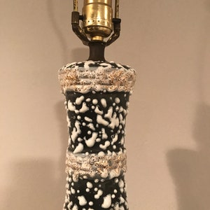 May include: A black and white ceramic table lamp with a textured glaze. The lamp has a gold-coloured socket and a white cord.