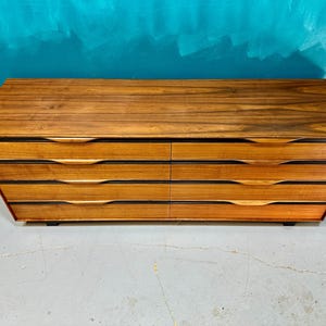 May include: A six-drawer dresser made of wood with a dark brown finish. The drawers have a curved, modern design and are set against a light gray background.
