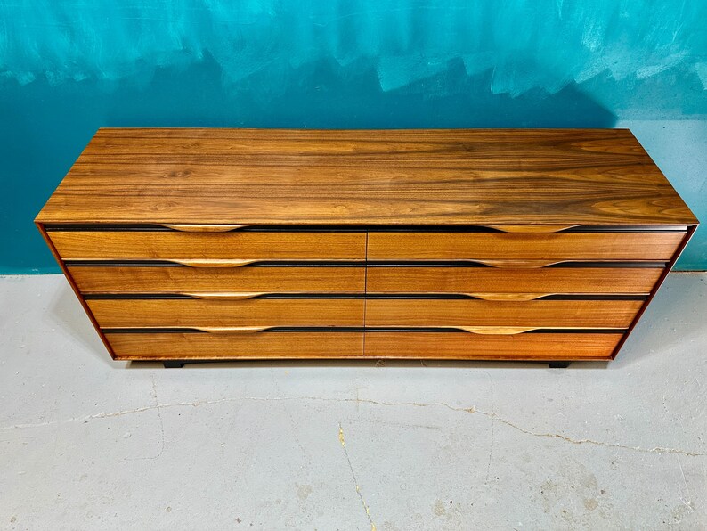 May include: A six-drawer dresser made of wood with a dark brown finish. The drawers have a curved, modern design and are set against a light gray background.