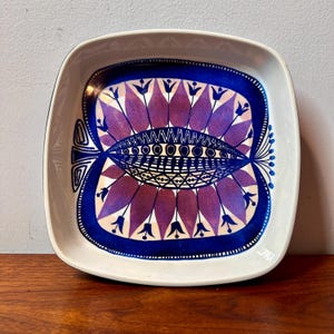 Puede incluir: Plato cuadrado de cerámica con borde blanco y un diseño simétrico vibrante. El interior presenta un motivo central de elementos florales estilizados morados y azules, con un borde azul oscuro. El plato está sobre una superficie de madera.