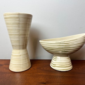 Peut inclure: Deux vases en céramique avec un motif rayé dans des tons beige et vert. Le vase de gauche est grand et fin, tandis que le vase de droite est plus court et plus large avec une ouverture évasée.