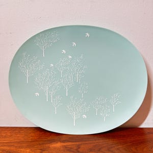 Op de afbeelding: Een grote, ovale serveerschaal in licht zeegroen. Witte, gestileerde bomen en vogels zijn op het oppervlak gedrukt. De schaal is waarschijnlijk gemaakt van keramiek of porselein en is geschikt voor het serveren van voedsel.