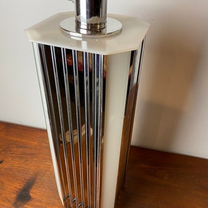 Op de afbeelding: Een chromen en witte tafellamp met een geometrisch ontwerp. De lamp heeft een cilindrische basis met verticale lijnen en een witte, matte glazen kap.