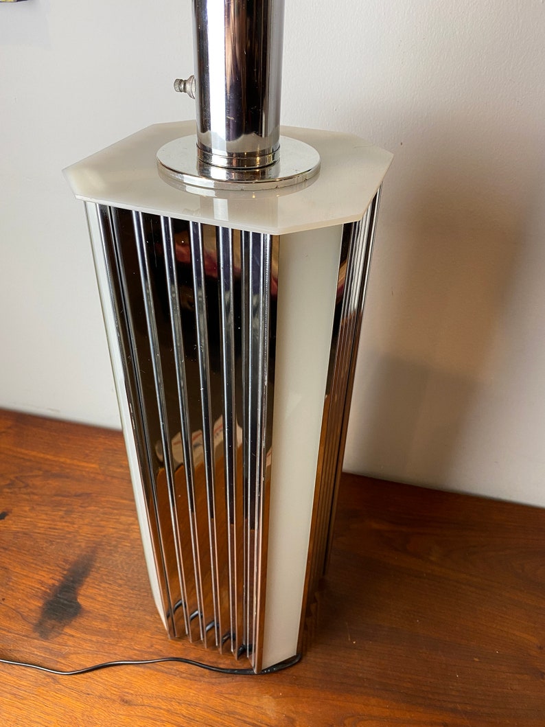 Op de afbeelding: Een chromen en witte tafellamp met een geometrisch ontwerp. De lamp heeft een cilindrische basis met verticale lijnen en een witte, matte glazen kap.