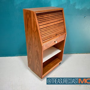 May include: A mid-century modern wooden cabinet with a roll-down shutter door and a drawer. The cabinet has two shelves and is on wheels. The cabinet is made of walnut wood and has a natural finish.
