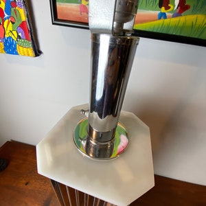 Op de afbeelding: Een chromen en witte tafellamp met een geometrische basis en een cilindrische kap. De lamp staat op een houten oppervlak.