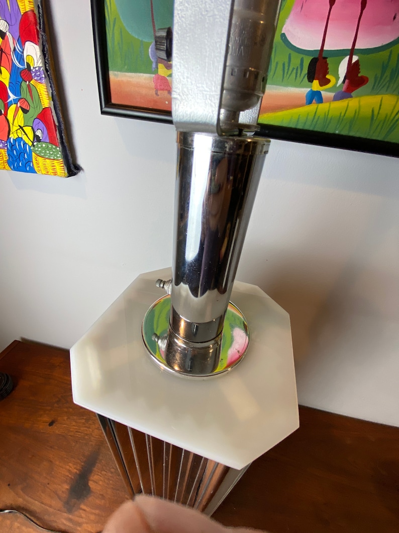 Op de afbeelding: Een chromen en witte tafellamp met een geometrische basis en een cilindrische kap. De lamp staat op een houten oppervlak.