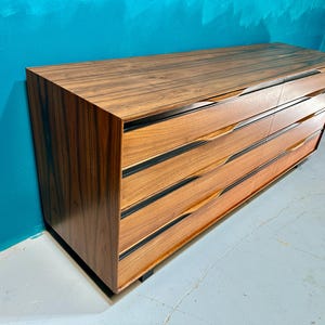 May include: A long, dark wood dresser with six drawers. The drawers have black trim and a unique curved design. The dresser is against a teal wall.