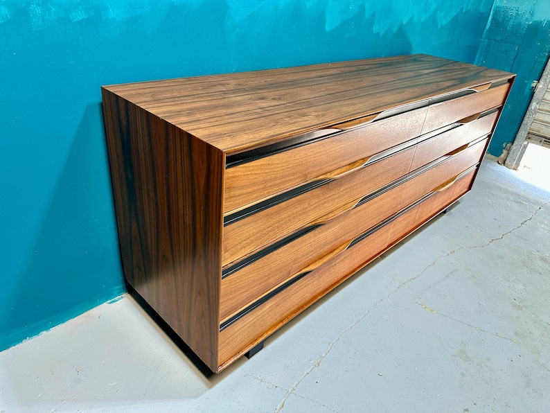May include: A long, dark wood dresser with six drawers. The drawers have black trim and a unique curved design. The dresser is against a teal wall.