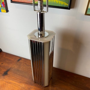 Op de afbeelding: Een chromen en witte tafellamp met een geometrische basis en een geribbeld ontwerp. De lamp heeft een witte kap en een chromen lampenkap.