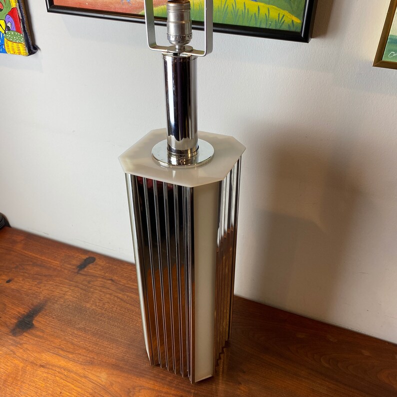 Op de afbeelding: Een chromen en witte tafellamp met een geometrische basis en een geribbeld ontwerp. De lamp heeft een witte kap en een chromen lampenkap.