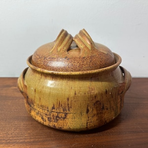 May include: A brown and yellow ceramic casserole dish with a lid. The lid has two handles and the dish has two handles on the sides. The dish has a rustic, handmade look.