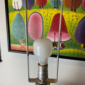 Op de afbeelding: Een zilveren tafellamp met een witte gloeilamp en een vierkante voet. De lamp staat voor een schilderij met een kleurrijke sc&egrave;ne met fruit en mensen.