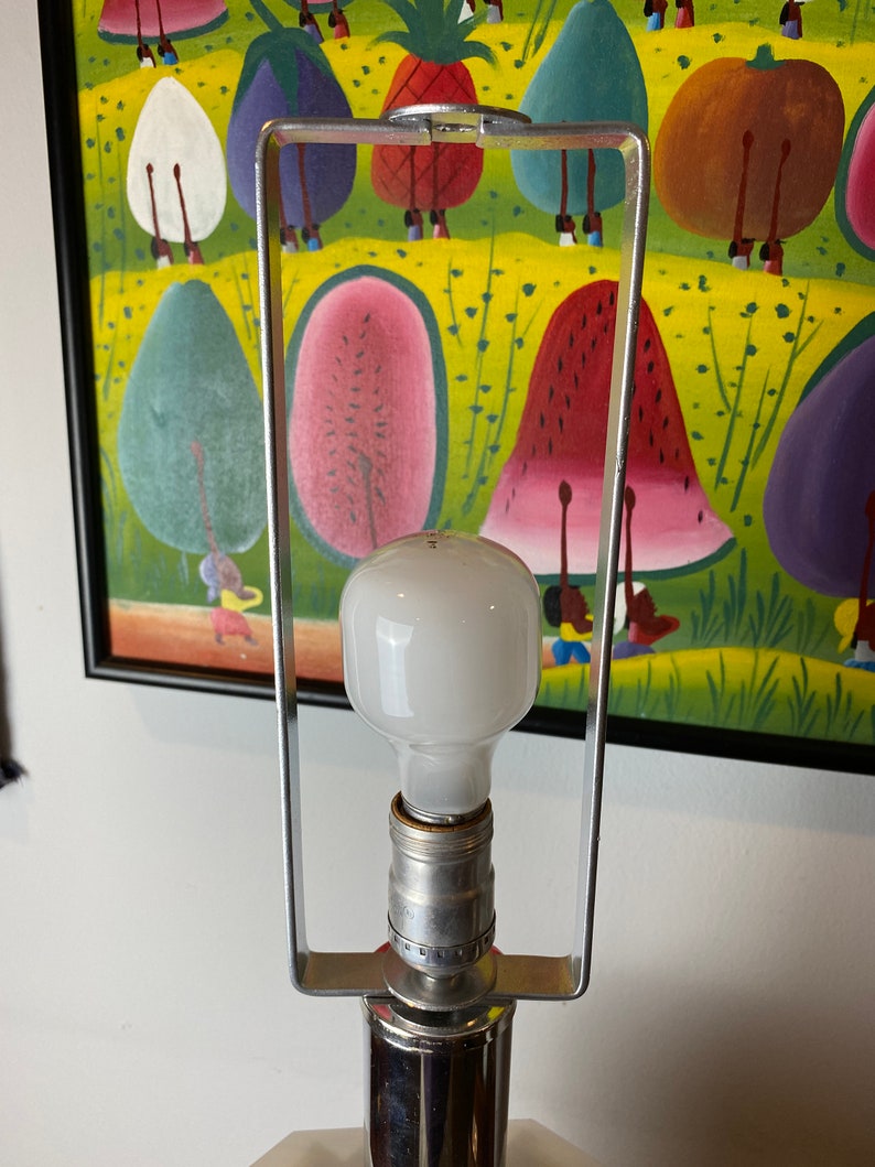 Op de afbeelding: Een zilveren tafellamp met een witte gloeilamp en een vierkante voet. De lamp staat voor een schilderij met een kleurrijke sc&egrave;ne met fruit en mensen.
