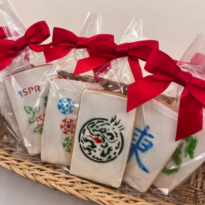 May include: Five white cookies decorated with colorful designs, including a dragon, Chinese characters, and a flower. The cookies are packaged in clear plastic bags tied with red bows.