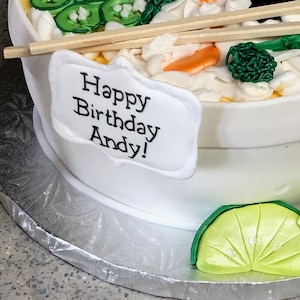 May include: A white bowl-shaped cake decorated to resemble ramen, featuring green scallions, orange carrot slices, and broccoli florets. Two wooden chopsticks rest on top. A white plaque on the side displays the black text Happy Birthday Andy!
