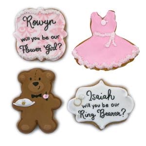 May include: Four decorated sugar cookies. Two cookies are shaped like signs with the text "Rowyn will you be our Flower Girl?" and "Isaiah will you be our Ring Bearer?". One cookie is shaped like a pink dress and the other is shaped like a brown teddy bear holding a small white plate.