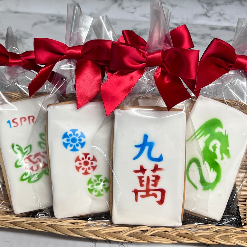 May include: Four decorated cookies in individual clear plastic bags tied with red bows. The cookies are shaped like mahjong tiles and feature different designs, including a green dragon, a red and blue flower, a green and red flower, and the Chinese characters for "Nine" and "Bamboo".