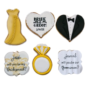 May include: Six decorated sugar cookies with wedding themes.  One cookie is shaped like a dress, another is a heart with the words "Bride & Groom" and the date "2/14/28", another is a heart shaped cookie with a tuxedo, another is a square cookie with the words "Jessi will you be my Bridesmaid?", another is a ring shaped cookie, and the last is a square cookie with the words "Jamal will you be our Groomsman?"