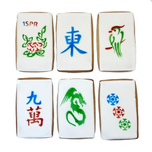 May include: Six decorated cookies shaped like mahjong tiles. The tiles feature various designs, including a green bird, a green dragon, a red flower, a blue Chinese character, and two red and blue Chinese characters. The tiles are decorated with green, red, and blue paint.