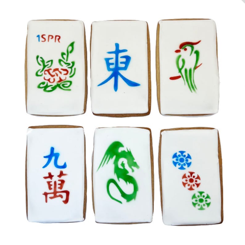 May include: Six decorated cookies shaped like mahjong tiles. The tiles feature various designs, including a green bird, a green dragon, a red flower, a blue Chinese character, and two red and blue Chinese characters. The tiles are decorated with green, red, and blue paint.
