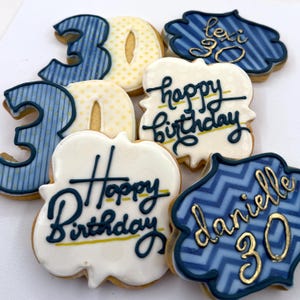 May include: A collection of decorated sugar cookies featuring two number 30 cookies with blue stripes and yellow dots, two white plaque cookies with dark blue 'happy birthday' text, and two blue plaque cookies with gold names and the number 30.
