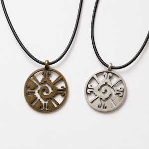 May include: Two circular pendants on black cords. One pendant is finished in an antique bronze tone, while the other is in a silver tone. Both feature an openwork design with a central spiral surrounded by radiating geometric shapes.