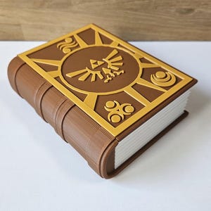 May include: A brown book with a gold embossed cover featuring the Triforce symbol and other intricate designs. The book is closed and appears to be made of a sturdy material.