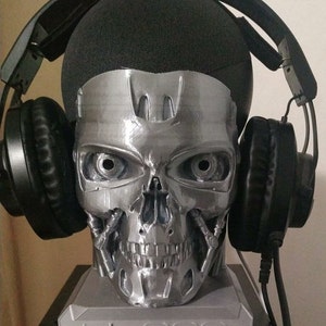 May include: A 3D printed silver Terminator T-800 skull headphone stand. The stand has a black headset resting on each ear. The stand is on a gray base with the text "T-800" printed on it.
