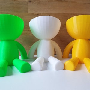 May include: Three colorful planters shaped like stylized figures with rounded limbs and heads. The planters are green, white, and yellow.