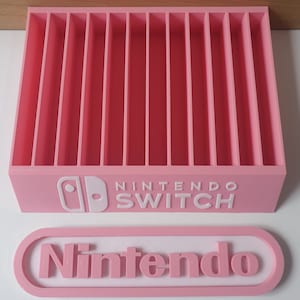 May include: Pink plastic storage case for Nintendo Switch game cartridges. The case has 12 slots and features the Nintendo Switch logo on the front. A separate pink plastic label with the word "Nintendo" is also included.