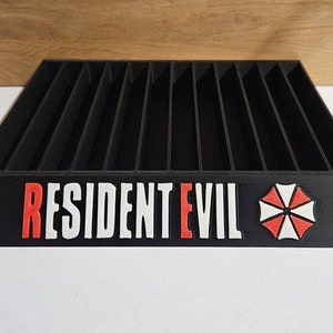 May include: Black plastic game case with slots for storing video game cartridges. The case has the words "RESIDENT EVIL" in white letters with a red "E" and a red and white umbrella logo.
